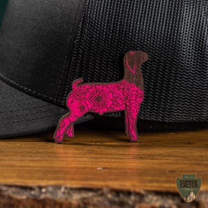 May include: A wooden dog-shaped pin with a floral design in pink and brown. The pin is placed on a wooden surface next to a black baseball cap. The pin has a detailed floral pattern.