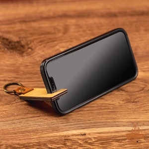 May include: A wooden phone stand with a keychain attached. The stand is made of light brown wood and has a black phone resting on it. The stand is on a wooden surface.