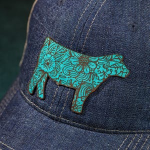 Puede incluir: Una gorra de béisbol de mezclilla con un diseño de silueta de vaca floral azul turquesa.