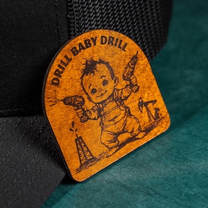 May include: A brown leather patch with a cartoon illustration of a baby holding two power drills. The text "DRILL BABY DRILL" is written in a circle around the baby.