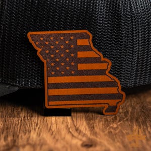 以下が含まれることがあります： ミズーリ州の形をした茶色の革製パッチ。アメリカ国旗のデザインが施されています。パッチは黒い背景と木製の表面に配置されています。国旗のデザインには星とストライプが含まれています。
