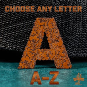 May include: A brown leather letter A patch featuring an intricate floral tooling pattern. The text CHOOSE ANY LETTER is displayed above, with A-Z and a Copper Cactus logo visible below. The patch is positioned against a black mesh background.