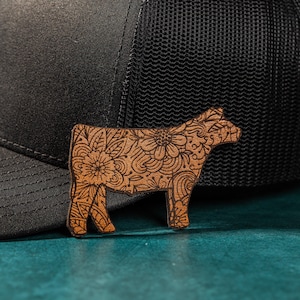 May include: A brown wooden cutout of a cow with a floral design. The cutout is sitting on top of a black and gray baseball cap.