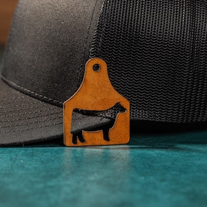 May include: A black and brown trucker hat with a leather cow tag attached. The tag has a silhouette of a cow.