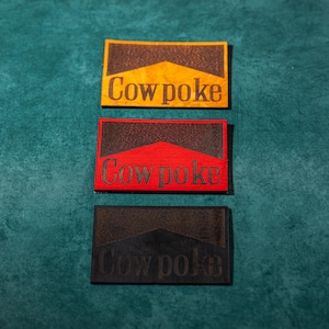 May include: Three leather patches with the text "Cowpoke" in black lettering. The patches are yellow, red, and black.