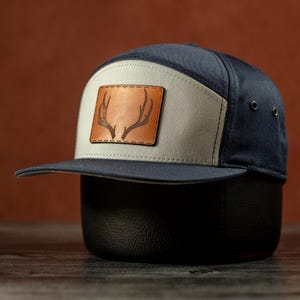 Può includere: Un cappello da baseball blu navy e beige con una toppa in pelle marrone che presenta un design a corna di cervo.