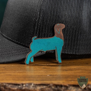May include: A wooden dog-shaped pin with a teal body and brown head, placed on a black trucker hat. The pin is on a wooden surface. The Copper Cactus logo is visible in the bottom right corner.
