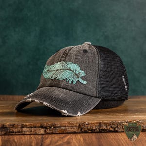 May include: A distressed black baseball cap with a black mesh back and a turquoise feather graphic on the front. The cap features a curved, frayed brim and a button on top. A small logo tag with the text Copper Cactus Collection is visible.