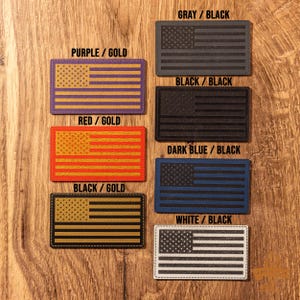 May include: Six different color combinations of American flag patches. The patches are made of leather and have a stitched border. The colors include gray, black, purple, gold, red, dark blue, and white.