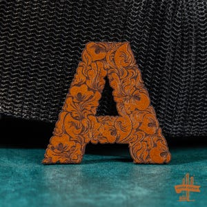May include: A brown leather letter A with a decorative floral pattern. The letter is on a black and teal background.
