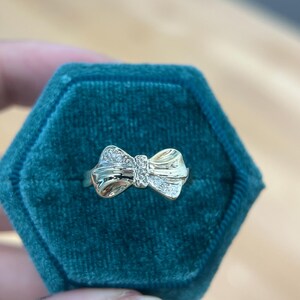 Può includere: Un anello a forma di fiocco dorato, con un gruppo centrale di piccole pietre preziose trasparenti. L'anello è presentato su uno sfondo di velluto verde acqua, che ne evidenzia il design delicato e la brillantezza. I dettagli del fiocco sono ben definiti.
