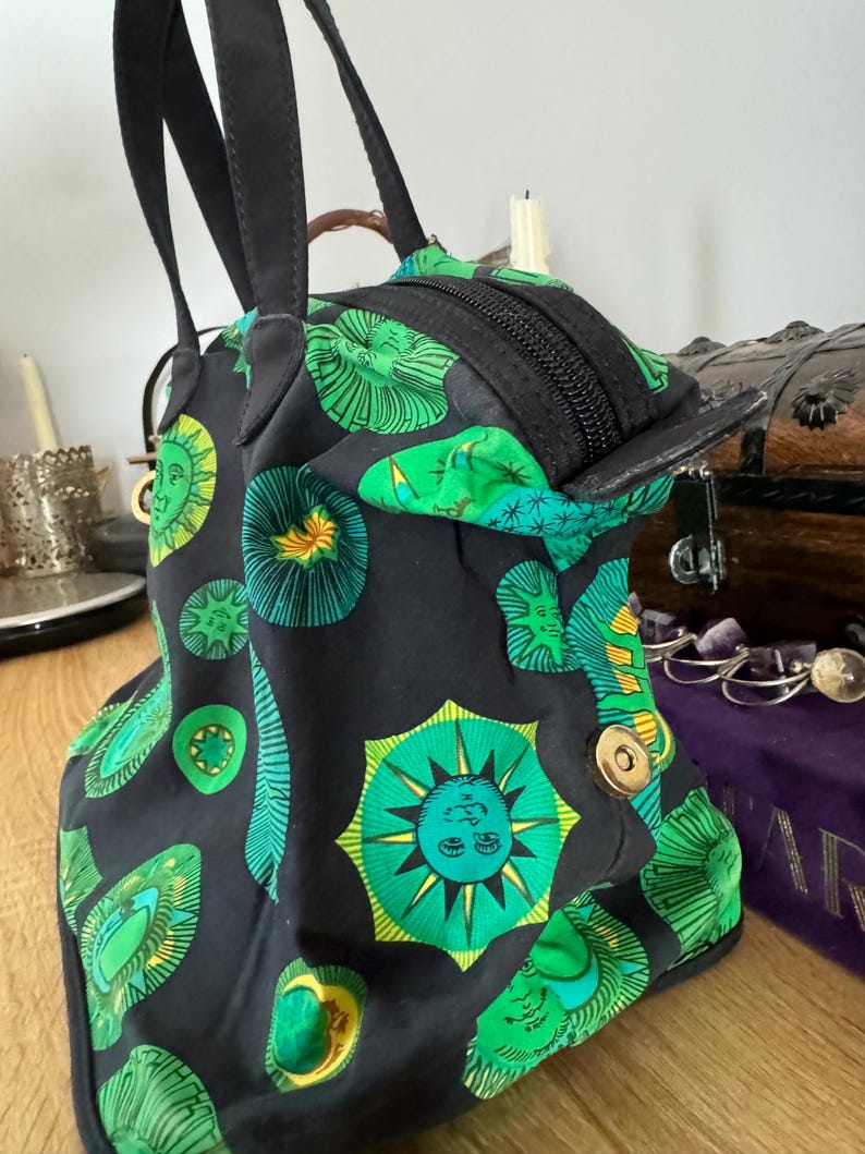 May include: A black tote bag with green and yellow sun and star designs. The bag has two black handles and a zipper closure.