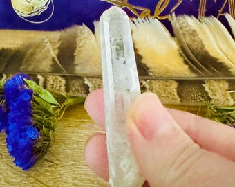 May include: A clear quartz crystal point with a slightly cloudy appearance. The crystal is about 5 inches long and is held in a hand.