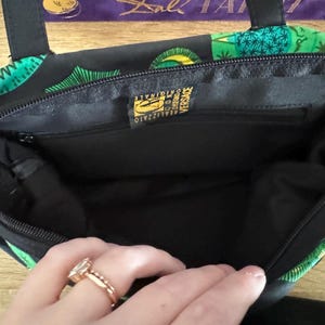 May include: Black handbag with a green and gold floral print. The inside of the bag is black and has a gold label that reads "Realizzato in Italia Versace Originale".