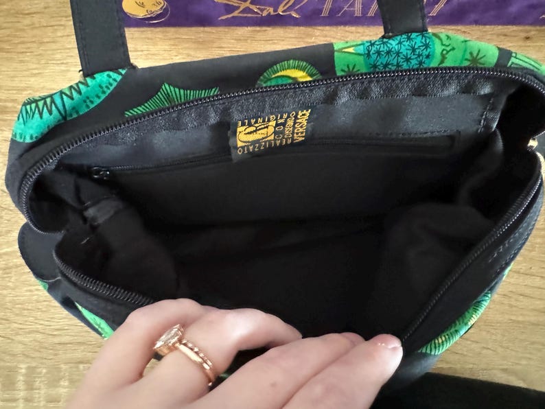 May include: Black handbag with a green and gold floral print. The inside of the bag is black and has a gold label that reads "Realizzato in Italia Versace Originale".