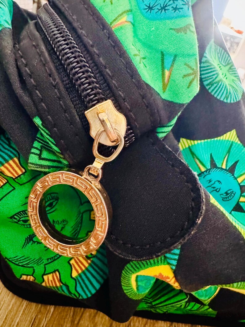 May include: A black zippered bag with a green and yellow patterned fabric. The zipper has a gold charm with a circular design.