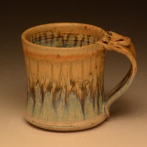 May include: A ceramic mug with a cylindrical body and a curved handle. The exterior features a glaze with a warm tan upper section that transitions into a blue and brown drip pattern toward the base. The interior rim shows a similar tan glaze.