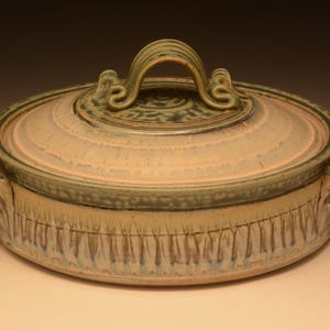 May include: A round ceramic casserole dish with a matching lid, featuring a beige base with vertical blue and green drip glaze patterns. The lid has a central arched handle, and the base includes two side handles with decorative scroll details.