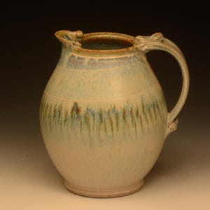May include: A ceramic pitcher with a rounded body and a curved handle. The surface features a cream-colored base glaze with vertical blue and brown drip patterns cascading from the rim. The pitcher has a wide opening and a small pouring spout.