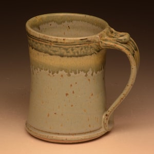May include: A handmade ceramic mug with a unique handle design. The mug features a light beige color with brown speckles and a gradient of yellow and green near the rim. The handle has a decorative, textured design.