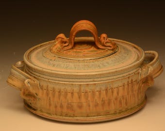 May include: A round ceramic casserole dish with a lid, featuring a warm tan and light blue glaze. The exterior has a textured, repeating vertical pattern, and the lid includes a prominent arched handle with decorative side accents.