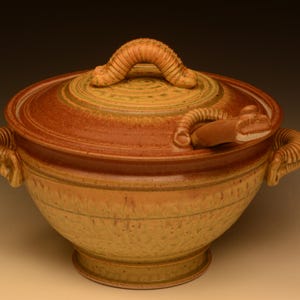 May include: A handcrafted ceramic lidded bowl in shades of brown and tan. The bowl features a textured surface, a decorative handle on the lid, and two side handles. The lid has a unique, curved handle. The bowl is suitable for serving.