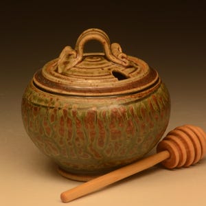 Puede incluir: Un tarro de miel de cerámica con tapa y un cucharón de miel de madera. El tarro es redondo con un esmalte moteado verde y marrón. La tapa tiene un asa y una pequeña abertura. El cucharón de miel es de color marrón claro.