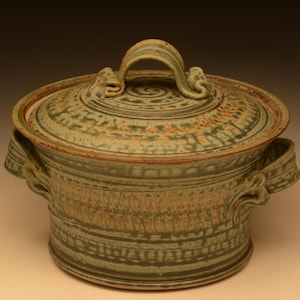 Op de afbeelding: Een groene en bruine keramische ovenschaal met deksel. De schaal heeft een ronde vorm, een deksel met handvat en twee zijhandvatten. Deksel en lichaam hebben decoratieve patronen. Geschikt om in te koken en te serveren.