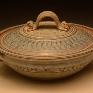 May include: A round ceramic casserole dish with a matching lid, featuring a speckled beige glaze with blue and pinkish-brown accents. The lid has a central arched handle and a decorative radial pattern, while the base includes two side handles.