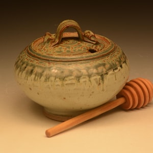 May include: A round ceramic honey pot with a lid featuring a handle and a small opening. The pot has a speckled cream base with green drip glaze accents around the top rim. A light brown wooden honey dipper with a grooved head rests beside the pot.