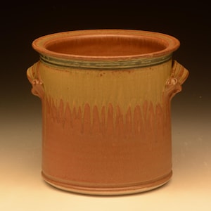 May include: A cylindrical ceramic crock featuring a wide rim and two side handles. The exterior displays a two-tone glaze with a warm terracotta base color transitioning into a lighter tan upper section, accented by a thin dark green band near the top.