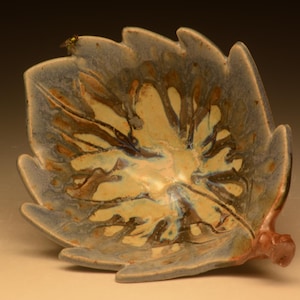 Op de afbeelding: Een keramische kom in de vorm van een blad met een glanzend glazuur in een mix van crème, bruine en blauwe kleuren. Het oppervlak heeft een gemarmerd, druipend patroon dat vanuit het midden naar de gekartelde randen straalt, met een steeltje.
