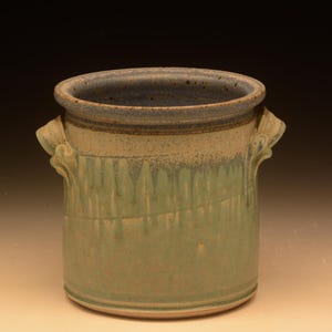 May include: A cylindrical ceramic crock featuring a light beige upper section and a sage green lower section with a vertical drip glaze pattern. The vessel includes two small, curved side handles and a dark blue-grey rim with speckled texture.