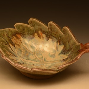 May include: A ceramic leaf-shaped bowl with a naturalistic design. The bowl features a light green interior with brown and tan accents, and a brown rim. The bowl is approximately 10 inches wide and 8 inches long.