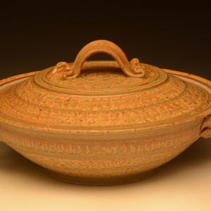Puede incluir: Un cuenco de cerámica hecho a mano con tapa en tonos beige y marrón. El cuenco tiene forma redondeada con tapa a juego y asa curvada. Los lados tienen asas decorativas y patrones texturizados.