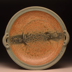 May include: A handmade ceramic serving platter with a speckled orange and tan surface, accented by a teal rim and handles. The center features a spiral design. The handles have a ribbed texture.