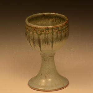 May include: A handmade ceramic goblet with a wide bowl and a flared base. The goblet is glazed in shades of green and brown, with a textured rim and a smooth, glossy finish. The goblet stands approximately 15 cm tall.