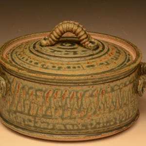 May include: A handcrafted ceramic casserole dish with a lid. The dish is a muted green with reddish-brown accents and a textured surface. It features a looped handle on the lid and two side handles. The dish measures approximately 10 inches in diameter.