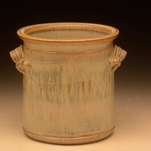 May include: A cylindrical ceramic crock featuring two side handles and a wide opening. The exterior surface displays a mottled glaze with vertical streaks of beige, cream, and soft sage green, finished with a warm brown rim and base edge.