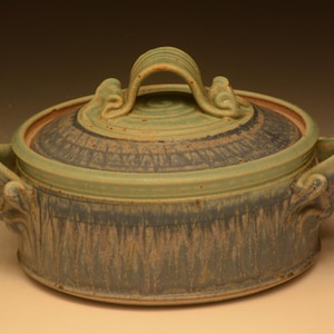 May include: A round ceramic casserole dish with a lid, featuring a sage green and slate blue glaze with vertical drip patterns. The lid has a central arched handle, and the base includes two side handles with decorative scroll-like details.