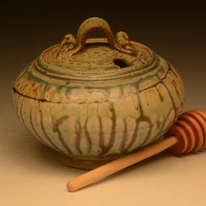 May include: A round ceramic honey pot with a lid featuring a looped handle and a small notch for a dipper. The pot has a mottled glaze in shades of cream, green, and brown with vertical drip patterns. A light-colored wooden honey dipper rests nearby.