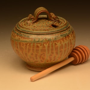 Peut inclure: Un pot à miel en céramique ronde avec un couvercle présentant un vernis texturé et tacheté dans des tons de beige, brun et vert. Le pot possède une petite ouverture pour un ustensile et est accompagné d'une cuillère à miel en bois clair.