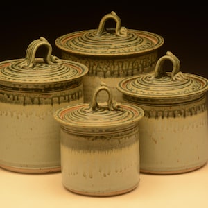 May include: A set of four cylindrical ceramic canisters with lids, each featuring a light cream base, a dark green drip glaze pattern around the upper rim, and a looped handle on the lid. The lids have a circular, swirling texture pattern.