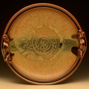 May include: A handmade ceramic serving plate with a spiral design in the center. The plate features a brown rim and handles, with a blend of green, orange, and brown colors. The plate is round and has a glossy finish.
