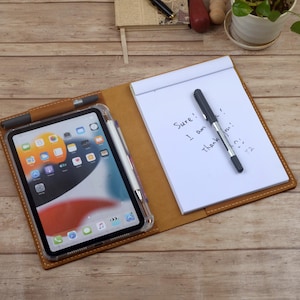May include: A tan leather folio organizer featuring a tablet in a clear case on the left side and a white notepad on the right. The notepad contains handwritten text that reads Sure! I am thank... T2. A black pen rests on the notepad.
