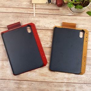 May include: Two tablet cases featuring a black interior shell with a camera cutout, attached to a leather side panel with punched holes for binding. One case has a deep red leather side, while the other has a tan leather side with visible stitching.