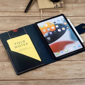Könnte beinhalten: Eine blaue Ledermappe mit einer Tasche für ein Notizbuch und einem Fach für ein Tablet. Die Mappe hat eine Klappe, die mit einem Druckknopf geschlossen wird. Das Tablet ist ein iPad mit einem weißen Apple Pencil daneben. Das Notizbuch ist ein Notizbuch der Marke Field Notes.