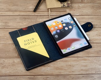 Może przedstawiać: Niebieski skórzany futerał typu folio z kieszenią na notes i miejscem na tablet. Futerał ma klapę zamykaną na zatrzask. Tablet to iPad z białym ołówkiem Apple Pencil obok. Notes to notes marki Field Notes.