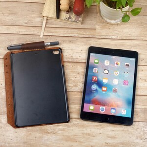 May include: A dark brown leather tablet case with a pen holder loop and a silver pen, positioned next to a black tablet displaying a colorful home screen with various app icons. The case features visible stitching along the edges and a flap closure.