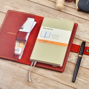 Può includere: Una copertina per taccuino in pelle rossa con un taccuino Moleskine Classic Collection a righe all'interno. La copertina del taccuino ha una patta con una tasca per carte o altri piccoli oggetti. Il taccuino è chiuso e ha un segnalibro a nastro. Una penna nera è appoggiata su una superficie di legno accanto al taccuino.